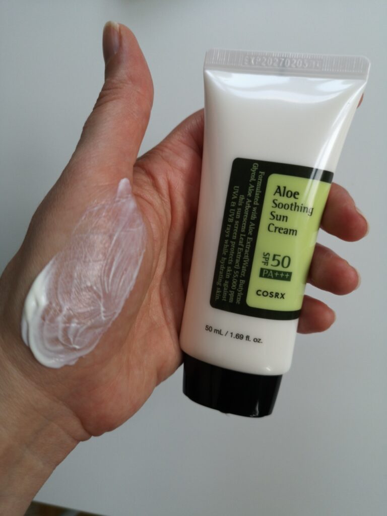 Jeg holder tuben med COSRX Aloe Soothing Sun Cream i hånden og har smurt litt av kremen på håndflaten for å vise konsistensen. Den er hvit og medium fyldig i konsistensen.